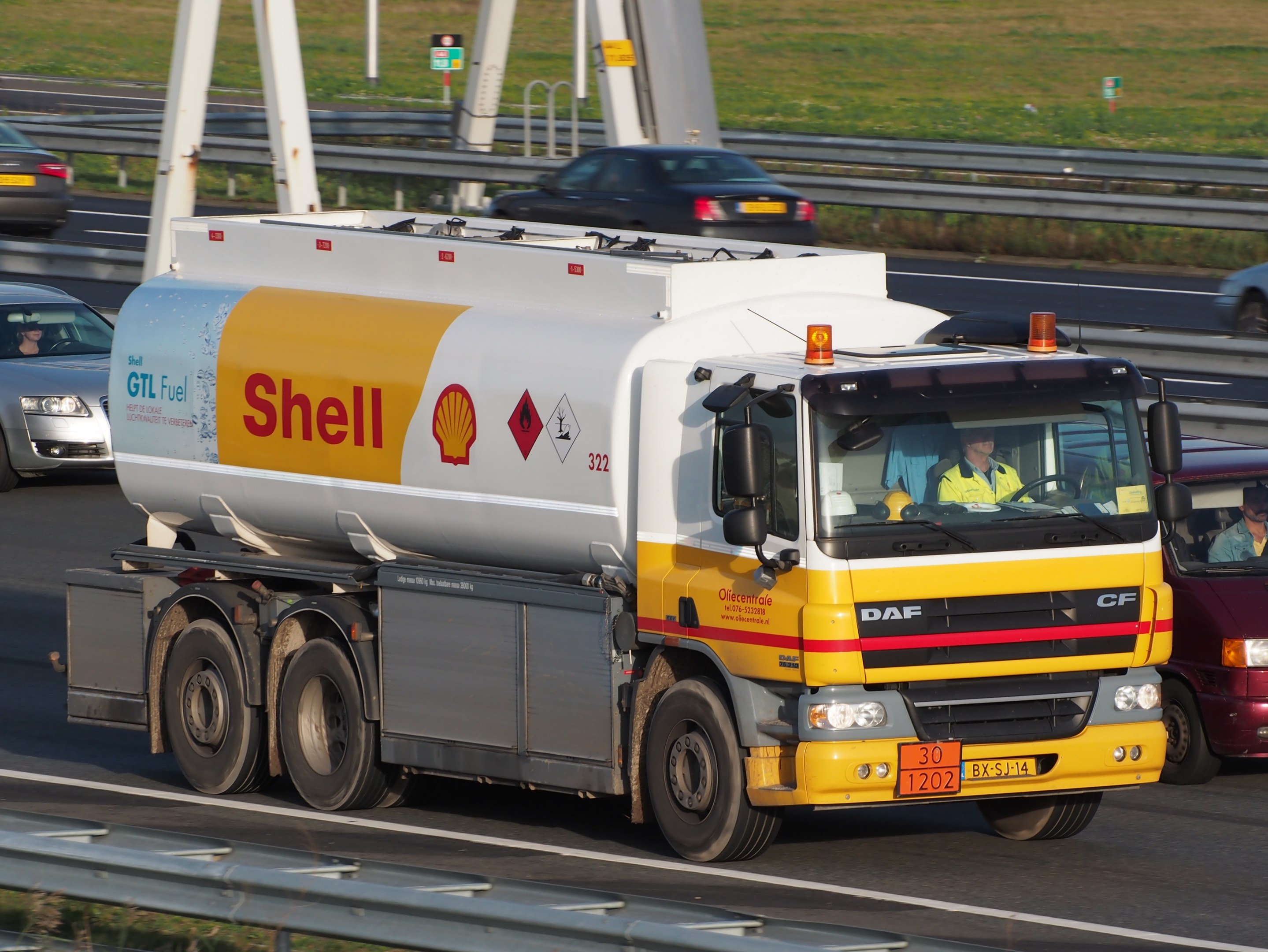 Ein Shell-Tanklastwagen auf einer Autobahn mit anderen Fahrzeugen, mit Menschen im Truck und Strommasten, Geländern, Beschilderungen und Gras im Hintergrund.