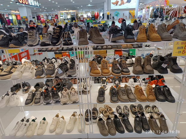 Ein Schuhgeschäfts-Innenraum mit verschiedenen Schuhen auf Ständern, Preisetiketten sichtbar, Menschen im Hintergrund, Kleiderauslagen, Deckenlampen, ein Bildschirm und Texttafeln.