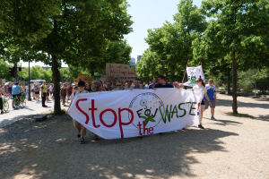 Eine Gruppe von Menschen, die eine 'Stop the Waste'-Tafel und verschiedene Plakate tragen und einige Fahrr├Ąder fahren, auf einer von B├Ąumen ges├Ąumten Stra├če, mit einigen Geb├Ąuden im Hintergrund unter einem klaren blauen Himmel.