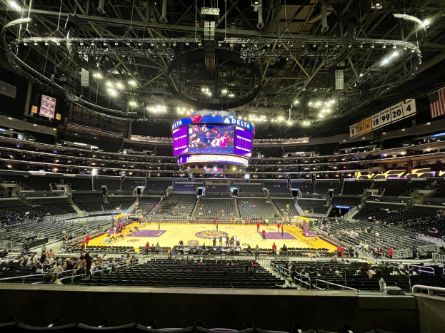 Eine große Indoor-Arena, das Staples Center, voller Zuschauer, die ein Basketballspiel verfolgen, mit Sitzbereichen, stehenden Zuschauern, einem prominenten Bildschirm oben, einer Flagge auf der rechten Seite, Scoreboards mit Text und Zahlen und Deckenstangen mit Beleuchtung, die eine lebendige Atmosphäre schaffen.