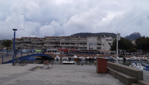 Ein Hafen mit Booten im Wasser, eine Br├╝cke mit Gel├Ândern, B├Ąnken, Laternen, B├Ąumen, Geb├Ąuden mit Fenstern, H├╝gel im Hintergrund und ein bew├Âlktes Himmel.