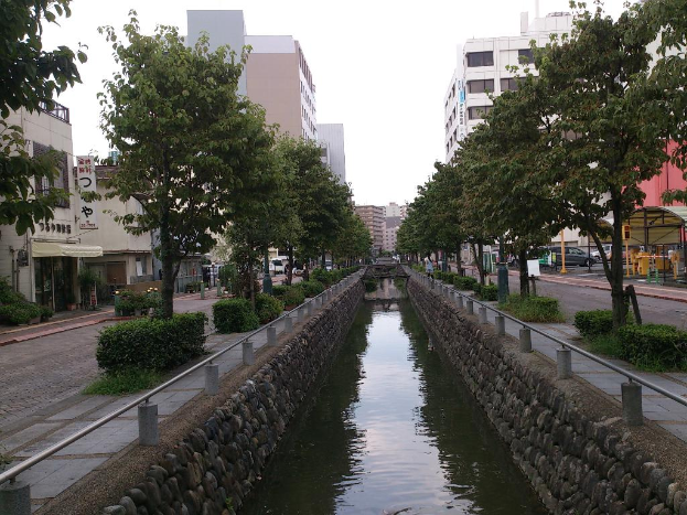 Ein Wasserkanal verläuft durch die Mitte des Bildes, flankiert von Hecken, Bäumen und einem Gehweg auf beiden Seiten, mit Straßen, Gebäuden und fahrenden Autos im Hintergrund unter einem Himmel.