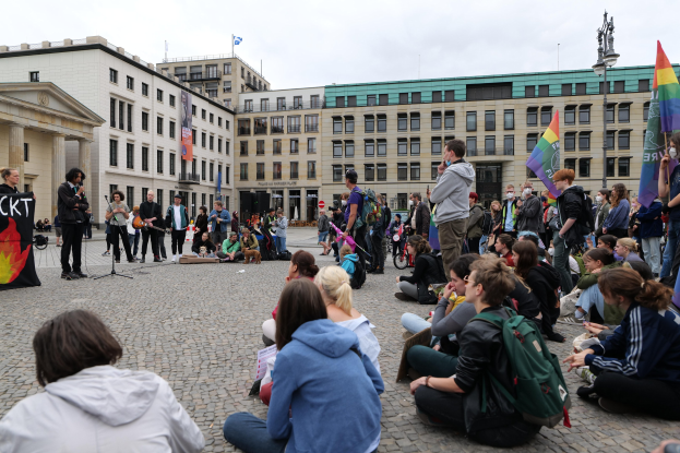 Eine Gruppe von Menschen, die auf dem Boden vor einer Menge sitzt, die Fahnen und Transparente hält, während einer Anti-Schwulen-Demo in Berlin, mit einer Statue, Gebäuden und einem Mikrofonständer zu sehen.