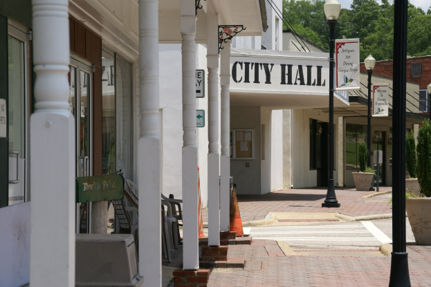 Ein von Gebäuden, Straßenlaternen, Straßenschildern, Kraftfahrzeugen, Verkehrskegeln, Stühlen, Topfpflanzen, Bäumen und einer bewölkten Himmel umgebenes Rathaus.