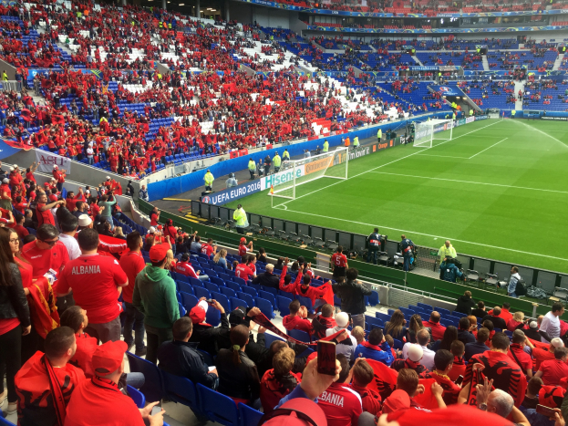 Ein volles Stadion mit sitzenden und stehenden Zuschauern, Bannern, einem Tor und Deckenbeleuchtung, während die Menge gespannt auf den Beginn des Fußballspiels wartet.