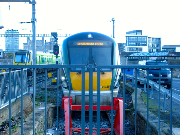 Zwei Züge auf parallelen Gleisen mit einem Zaun im Vordergrund, einem Auto auf der Straße rechts, einer Signalleuchte auf einem Pfahl und Gebäuden im Hintergrund unter einem Himmel.