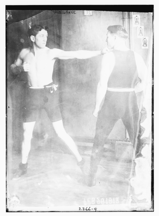 Schwarzes und weißes Foto von zwei Boxern in einem Ring mit Plakaten an der Hintergrundwand und Text unten.