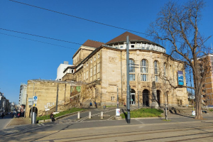Theater Kreiburg in Berlin, Deutschland, ein großes Gebäude mit zahlreichen Fenstern, umgeben von Straßenmasten, Straßenlaternen, Strommasten, Stromkabeln, Kraftfahrzeugen, Fußgängern, Bäumen und einem bewölkten Himmel.