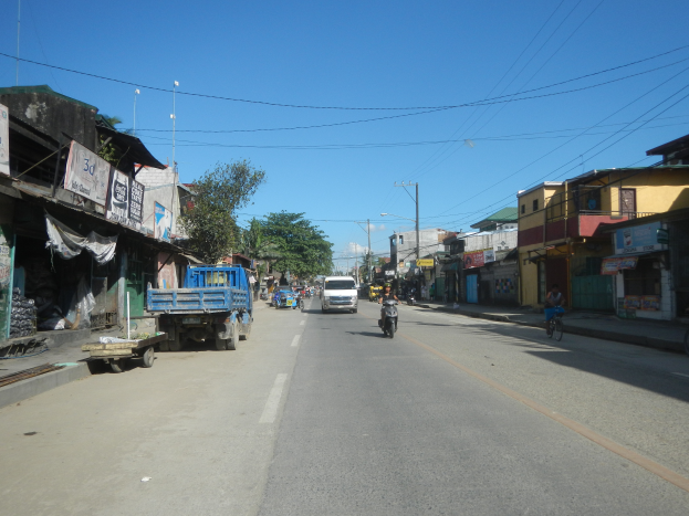 Eine Stadtstraße mit fahrenden Fahrzeugen, ein Radfahrer auf der rechten Seite, Gebäude auf beiden Seiten, Bäume im Hintergrund und ein klarer blauer Himmel.
