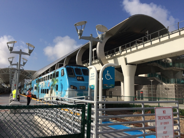 Ein paar Menschen auf einem linken Bahnsteig mit einem Zug auf der rechten Seite, eine Brücke, ein Zaun und mehrere Pfosten.