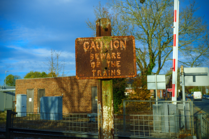Vorsichtsschild an einem Bahnübergang-Zaun neben Bäumen, Pfählen, einem Gebäude, Containern, Fahrzeugen und einem bewölkten Himmel.