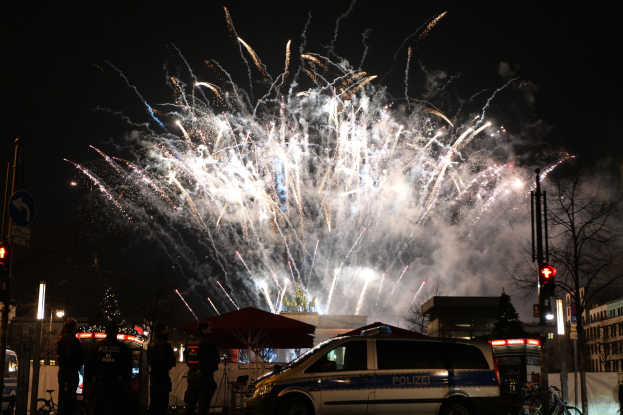 Polizeiauto vor einer Menschenmenge bei einer Silvesterfeier in Berlin geparkt, mit farbigen Feuerwerken am Himmel.