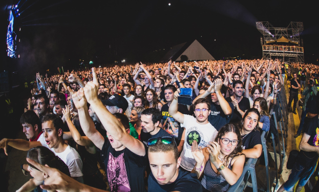Eine große Menschenmenge auf einem Musikfestival mit Menschen, die ihre Hände heben, einige sitzen und andere stehen, begrenzt von einem Metallzaun auf der rechten Seite, mit Gebäuden und Lichtern im Hintergrund unter einem dunklen Himmel.