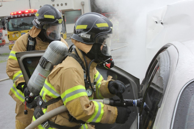 Feuerwehrleute in Schutzausrüstung verwenden einen Schlauch, um ein brennendes Auto zu löschen, mit Rauch und anderen Fahrzeugen und einem Gebäude im Hintergrund.