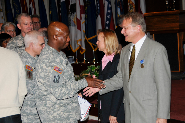 Eine Gruppe von Menschen, die mit zwei Personen Handschlag geben, Flaggen im Hintergrund, ein Podium mit Mikrofon und eine Topfpflanze in der Nähe.