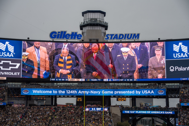 Eine große Menge Menschen in einem Stadion schaut ein Football-Spiel, mit einem zentralen Bildschirm mit Bildern und Text, Schildern mit Text, Zuschauern in den Rängen, einem Bogen, Bäumen und einem klaren blauen Himmel im Hintergrund.