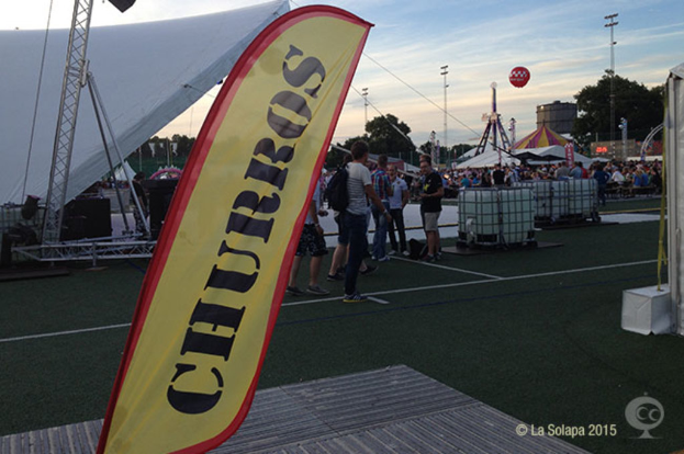 Ein lebhaftes Karnevalsszenario mit Zelten, einem zentralen Mast, Menschen, Bäumen, Strommasten, Straßenbahnen auf der Straße und einem Eisenständer links.
