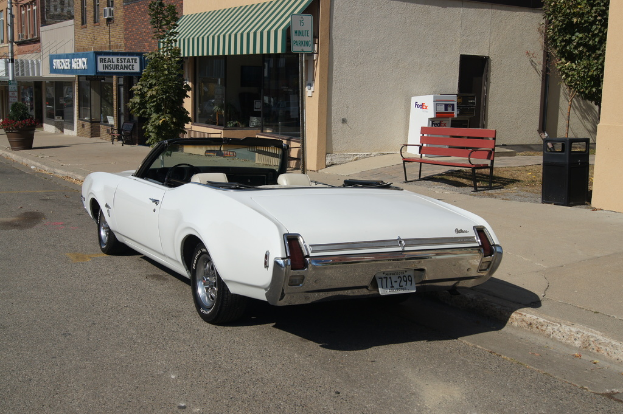Ein weißer Wagen mit der Nummer 771-299 steht auf einer Straße, rechts daneben eine Bank, im Hintergrund ein Gebäude und ein Ladengeschäft namens "Immobilienversicherung".