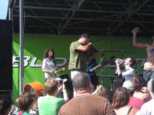 Eine Gruppe von Musikern, die verschiedene Instrumente auf einer Bühne spielen, während das Publikum vor einem Podium mit einem grünen Banner im Hintergrund steht.