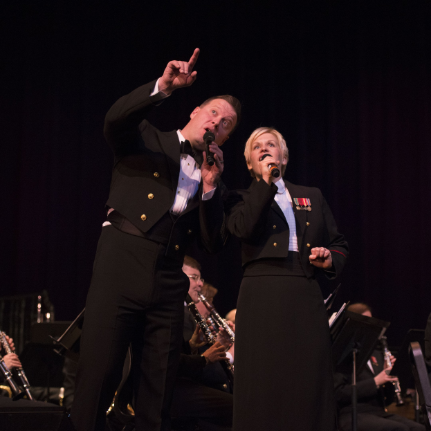 Ein Mann und eine Frau in Smokings stehen auf einer Bühne und halten Mikrofone, während eine Gruppe von Musikern hinter ihnen in einer dunklen Umgebung sitzt.