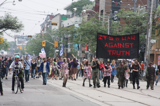 Eine Gruppe von Menschen marschiert auf einer Straße und hält Fahnen und Transparente hoch, mit einem Radfahrer in der Nähe, Laternenpfählen, Stromkabeln, Ampeln, Schildern, Bäumen und Gebäuden im Hintergrund.