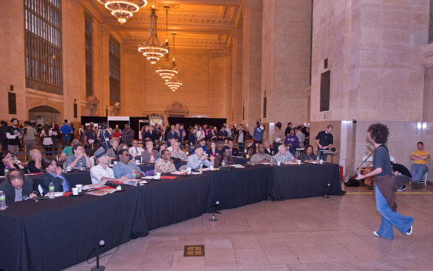 Eine Konzertszene mit einer Menge Menschen, einem Mann, der rechts Gitarre spielt und singt, einer Wand und Fenstern im Hintergrund, Lichtern oben und einem Boden unten.