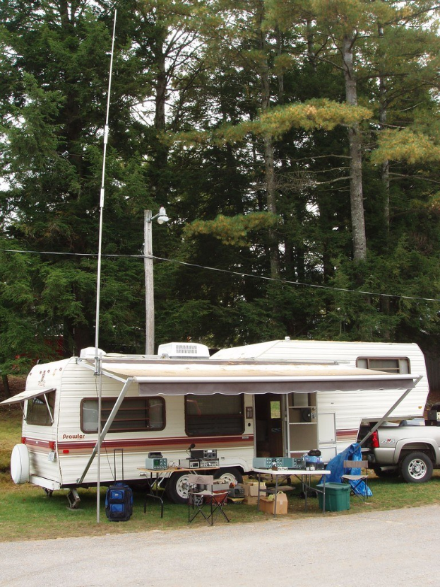 Wohnwagen mit Fenstern und Türen, Tischen, Kisten und einem Einkaufswagen davor, Gegenstände auf dem Tisch, eine Straße vorne, Bäume hinten und einem Laternenmast.