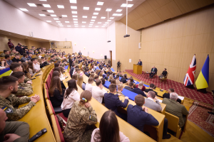 Eine große Gruppe von Menschen sitzt an Tischen in einem Raum mit Flaggen auf der rechten Seite, Papieren und Gegenständen auf den Tischen, einigen Individuen, die an einem Podium mit Mikrofon stehen, sowie Deckenleuchten und einem Projektor, was auf eine Militärakademie hindeutet.