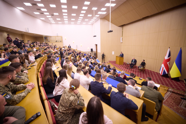 Eine große Gruppe von Menschen sitzt an Tischen in einem Raum mit Flaggen auf der rechten Seite, Papieren und Gegenständen auf den Tischen, einigen Individuen, die an einem Podium mit Mikrofon stehen, sowie Deckenleuchten und einem Projektor, was auf eine Militärakademie hindeutet.