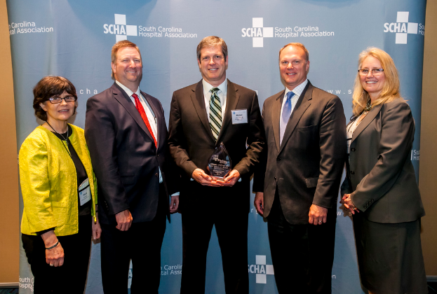 Eine Gruppe von Menschen, die meisten tragen Blazer, mit einer Person in einer gelben Jacke und zwei mit Brille, vor einem blauen Banner und einer gelben Wand.