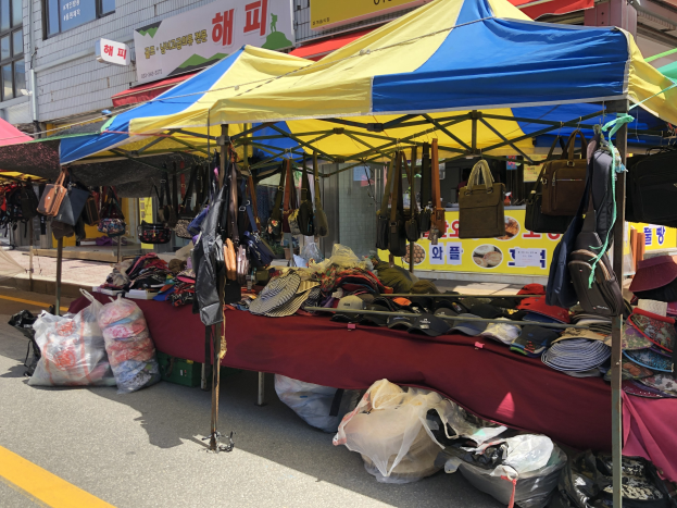 Ein Markt mit Zelten, die Taschen, Hüte und andere Gegenstände präsentieren, neben Gebäuden mit Fenstern und Texttafeln.