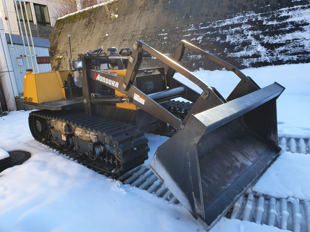 Ein gelb-schwarzer Radlader steht im Schnee vor einem Gebäude mit Bäumen und einem klaren Himmel.
