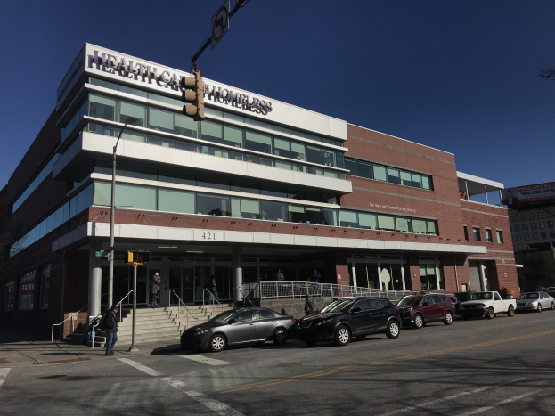Großes Gebäude, das als das New Yorker Gesundheitszentrum für Obdachlose gekennzeichnet ist, mit parkenden Autos davor, umgeben von Straßeninfrastruktur, Fußgängern auf dem Bürgersteig und Bäumen unter einem klaren blauen Himmel.