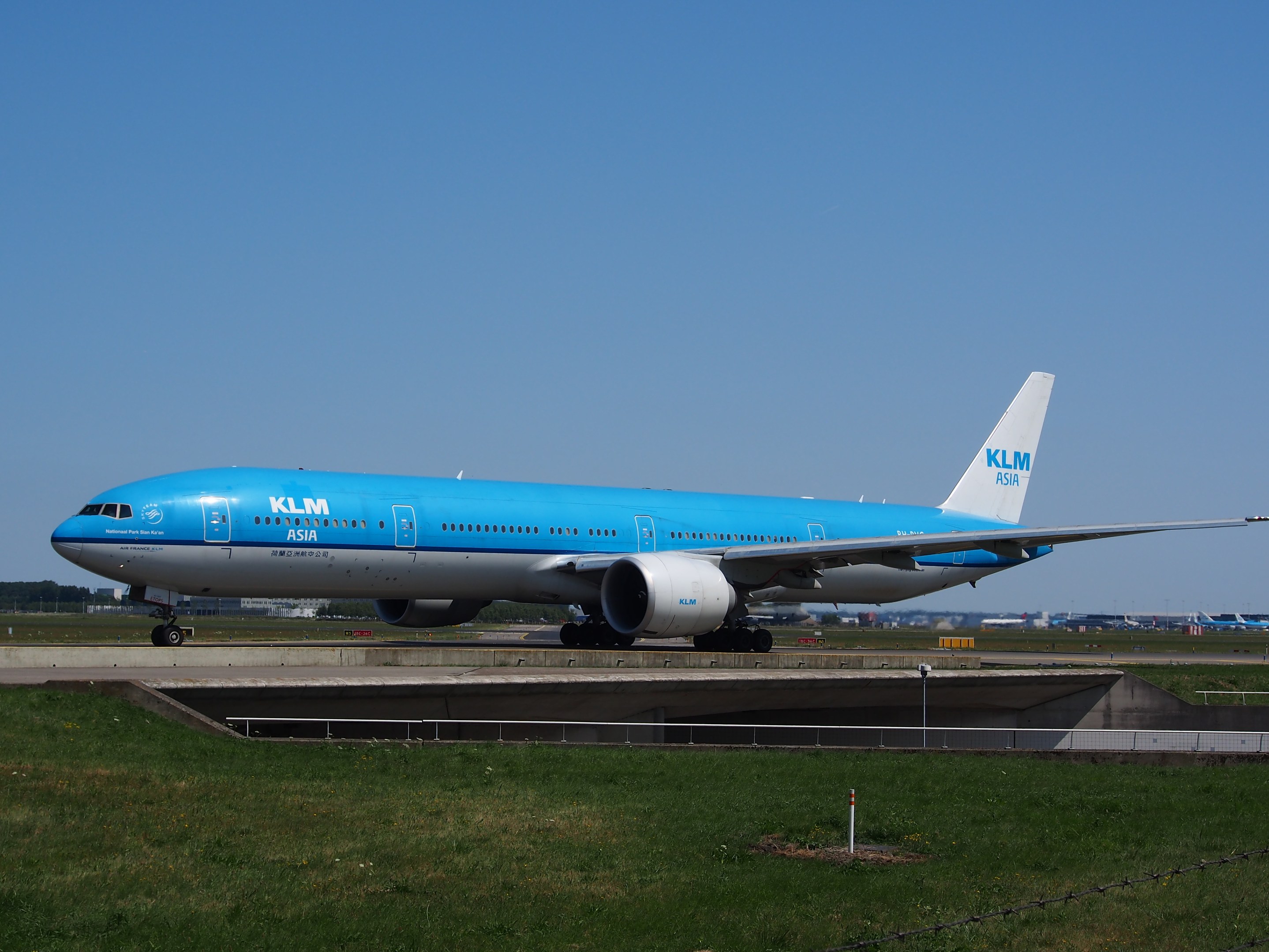 Ein KLM Boeing 777-300ER auf der Rollbahn am Amsterdam Airport, mit Gras, einem Zaun mit Pfählen, Gebäuden, Bäumen und einem klaren blauen Himmel im Hintergrund.