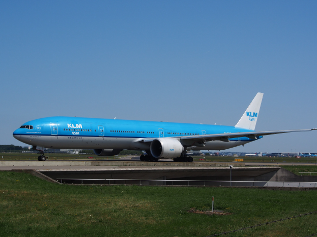 Ein KLM Boeing 777-300ER auf der Rollbahn am Amsterdam Airport, mit Gras, einem Zaun mit Pfählen, Gebäuden, Bäumen und einem klaren blauen Himmel im Hintergrund.
