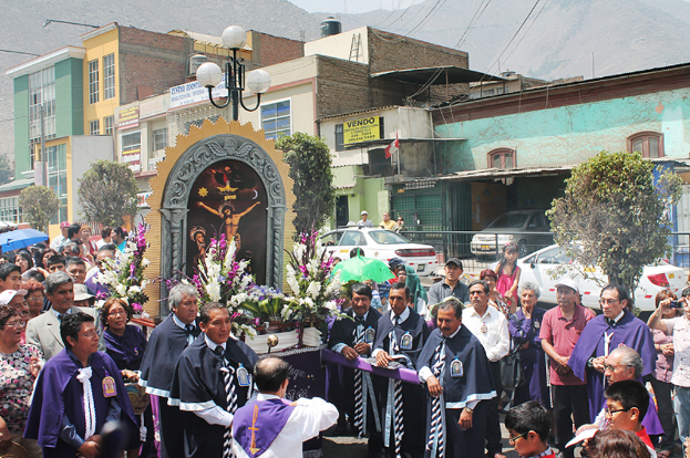 Menschen auf einer Straße mit einem Kreuz und Blumensträußen im Vordergrund, Autos in der Mitte und Gebäuden, Bäumen und Straßenlaternen im Hintergrund.