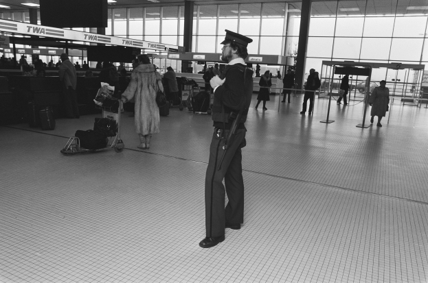 Polizeibeamter in Uniform mit einer Waffe in einem Flughafen-Terminal umgeben von Menschen und Gepäck mit Glaswänden und Informationsanzeigen im Hintergrund.