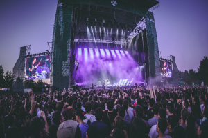 Eine große Menge Menschen auf einem Musikfestival schaut auf einer beleuchteten Bühne mit Instrumenten und Bildschirmen, die vor einem Gebäude, Bäumen und einem klaren blauen Himmel aufgebaut sind.