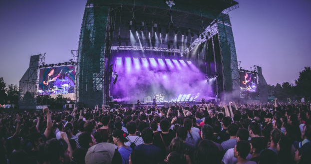 Eine große Menge Menschen auf einem Musikfestival schaut auf einer beleuchteten Bühne mit Instrumenten und Bildschirmen, die vor einem Gebäude, Bäumen und einem klaren blauen Himmel aufgebaut sind.