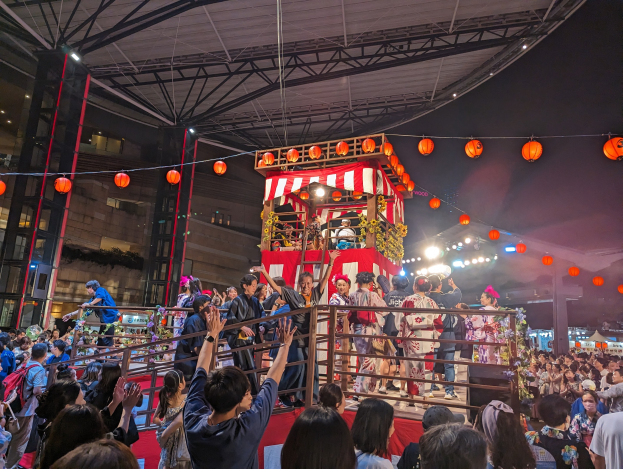 Eine Menschenmenge auf einer beleuchteten Bühne mit Laternen, einige halten Kameras, umgeben von einem Geländer, mit Bäumen und einem klaren blauen Himmel im Hintergrund, wahrscheinlich während eines japanischen Festivals.