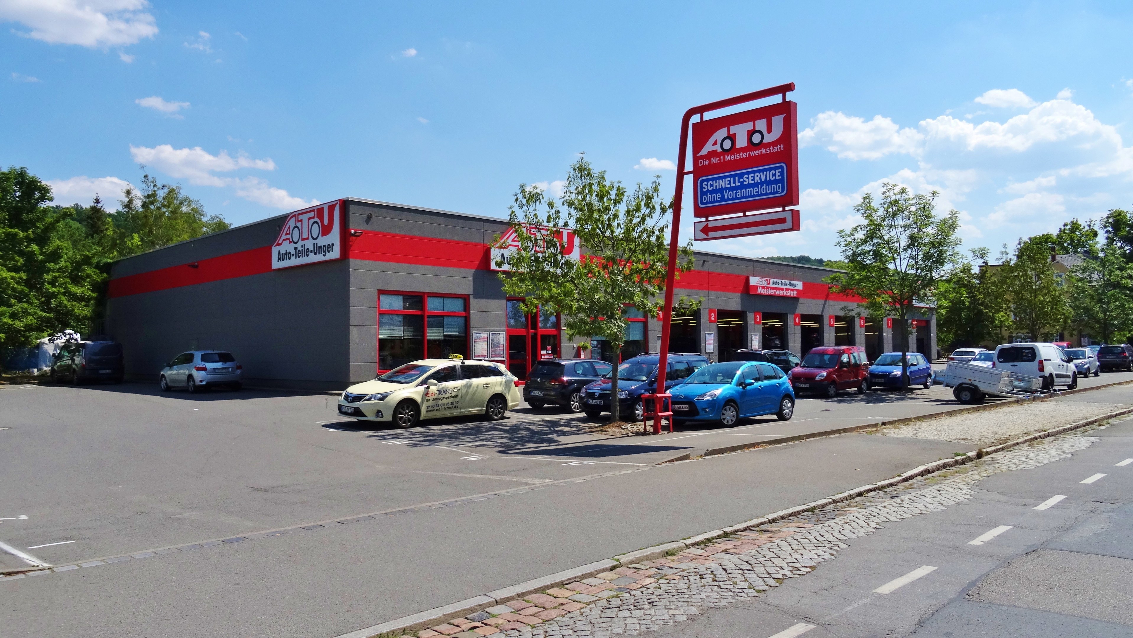 Ein Autohaus mit mehreren geparkten Fahrzeugen vorne, umgeben von Gebäuden, Bäumen, Strommasten, Schildern und einer Straße unter einem bewölkten Himmel.