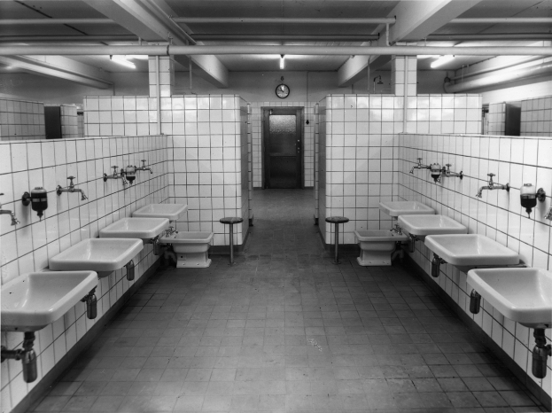 Ein Schwarz-Weiß-Foto einer öffentlichen Toilette mit Waschbecken, Wasserhähnen, Hockern, einer Wanduhr und Deckenleuchten.
