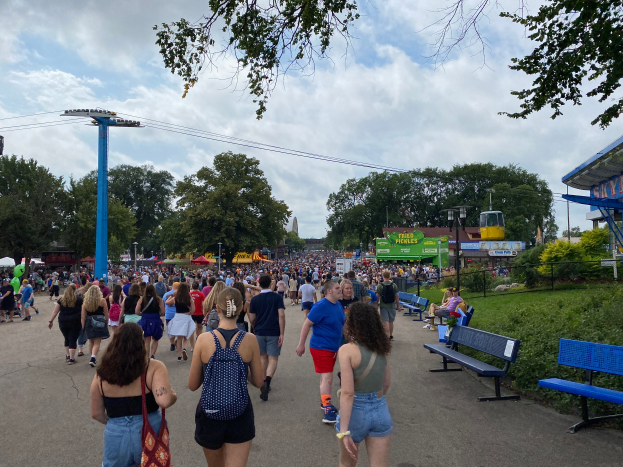 Eine Menschenmenge, die eine Straße entlanggeht, gesäumt von Bänken, Pflanzen, Gras, Bäumen, Pfählen, Laternen, Drähten und einem Schuppen, wobei einige Taschen tragen, unter einem bewölkten Himmel.
