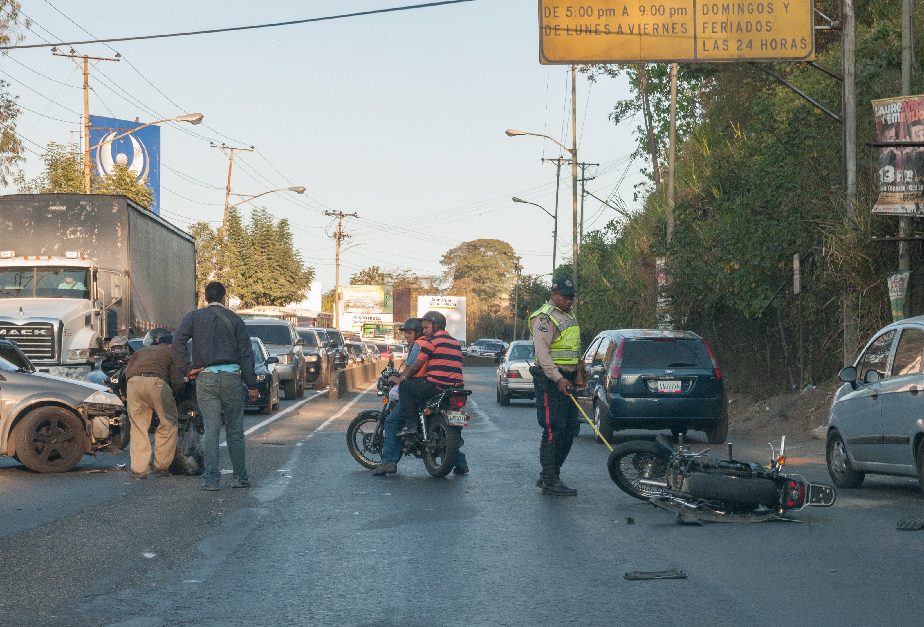 Gruppe von Menschen um ein verunglücktes Motorrad auf der Stra├če mit mehreren Fahrzeugen, B├Ąumen, Masten, Lichtern, Schildern und Himmel im Hintergrund.