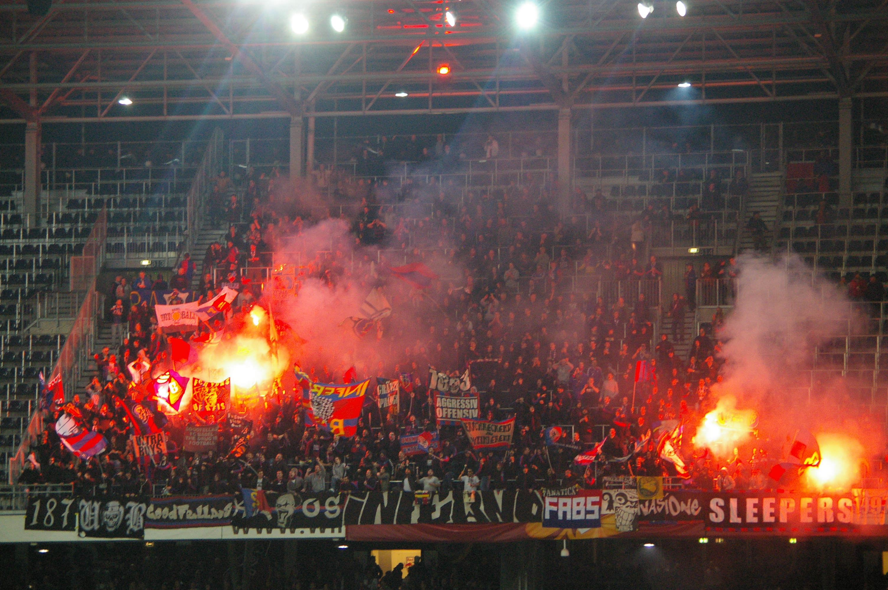 Eine große Menschenmenge in einem Stadion hält Fahnen und Banner, mit Leuchtraketen, die Rauch erzeugen, unter einer Decke mit Deckenlampen und Metallrahmen.