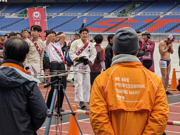 Eine Gruppe von Menschen auf einer Bahn mit einer Kamera auf einem Stativ im Vordergrund, Stühle, ein Banner und Lichter im Hintergrund.