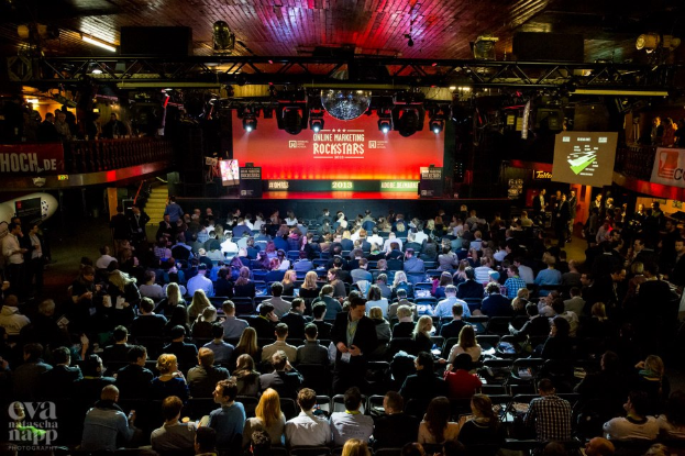 Eine große Menschenmenge sitzt auf Stühlen vor einer Bühne, die von Scheinwerfern beleuchtet und von Geländern umgeben ist. Auf der Bühne befinden sich Bildschirme und Banner mit Text, und in der linken unteren Ecke befindet sich ein Wasserzeichen, das eine Atmosphäre von Aufregung und Erwartung schafft.