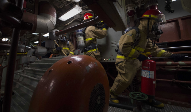 Feuerwehrleute in Schutzausrüstung arbeiten an einem Feuerhydranten in einem Raum mit Rohren, Stangen und Beleuchtung.