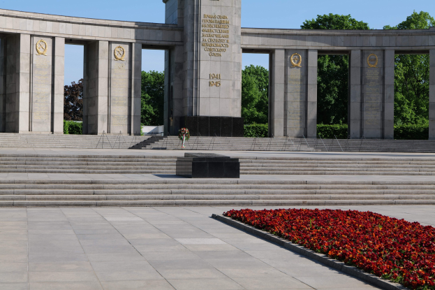 Ein steinernes Denkmal in einem Park, das mit Text beschriftet ist und von dem Stufen hinaufführen, umgeben von roten Blumen, Bäumen und einem klaren blauen Himmel.