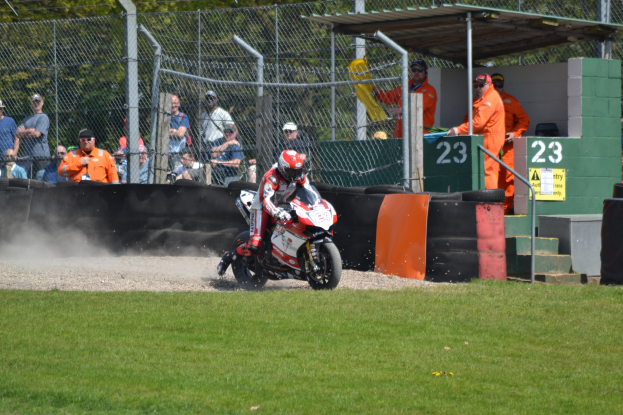 Eine Person in Helm und Handschuhen auf einem Motorrad auf einem grasbewachsenen Feld, mit Zuschauern in Mützen und Schutzbrillen, einem eingezäunten Bereich, Treppen mit Geländern und einem nummerierten Schuppen im Hintergrund.